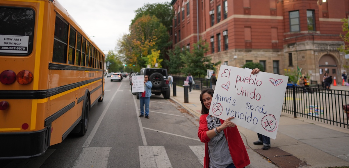 A teacher holds a sign between a school bus and the front of a school, with students visible in the background