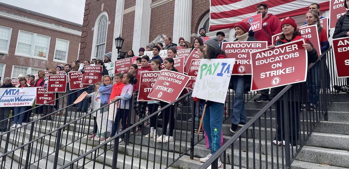 A large group of teachers stand in the steps of a large brick building holding red signs that say “Andover Educators on Strike”