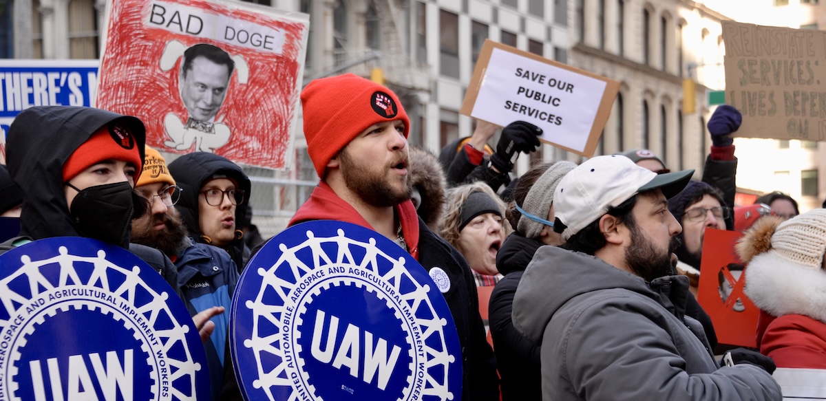 A group of 20 protesters with UAW and Save Our Services signs, many are yelling