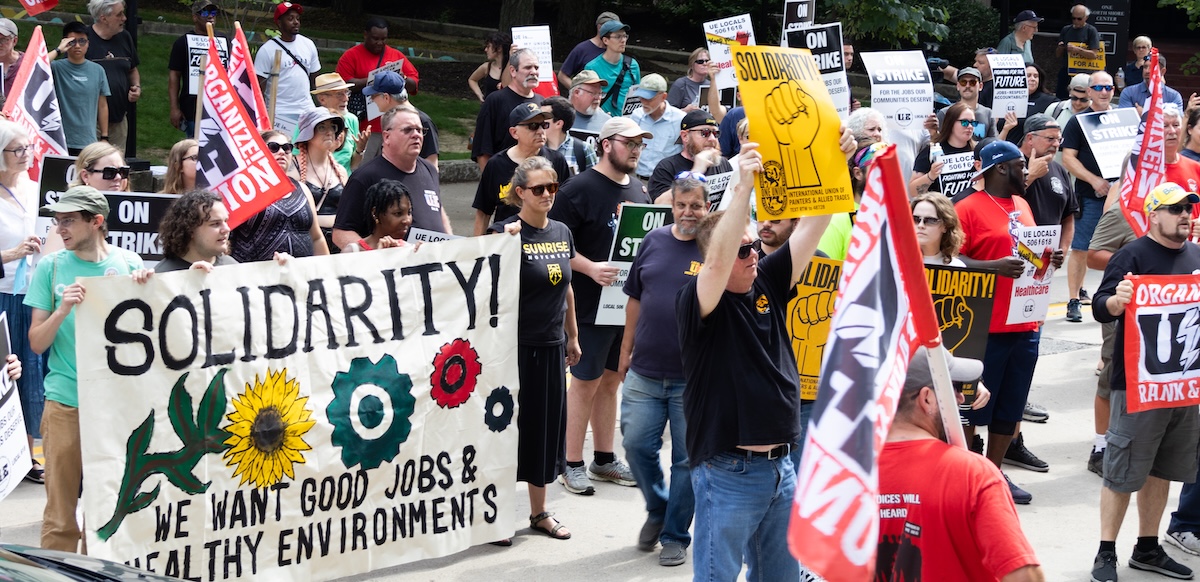 A rally with 30 or so people with a mixture of colorful signs, including Painters Union and a “We Want Good Jobs and Health Environments” banner