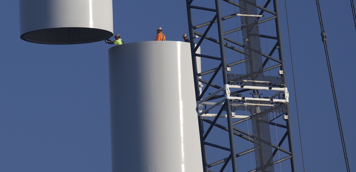 Workers join the top piece of a giant wind turbine to the bottom. They are tiny against its hugeness.