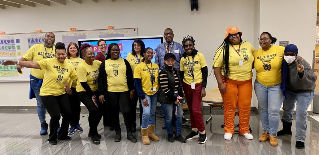 Fourteen workers, mostly Black and mostly women, pose smiling in a school classroom. Most are wearing yellow "Baltimore Teachers Union" T-shirts.