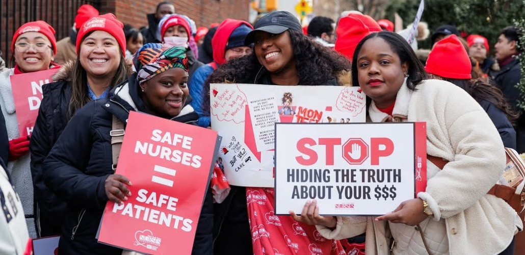Three Black women nurses pose for camera; two are smiling, one looks determined. Two hold printed NYSNA signs: "Safe nurses = safe patients" and "STOP hiding the truth about your $$$" and the other holds a handwritten sign, partly covered, long text. Lots of red NYSNA hats are visible in the dense, upbeat crowd behind them.