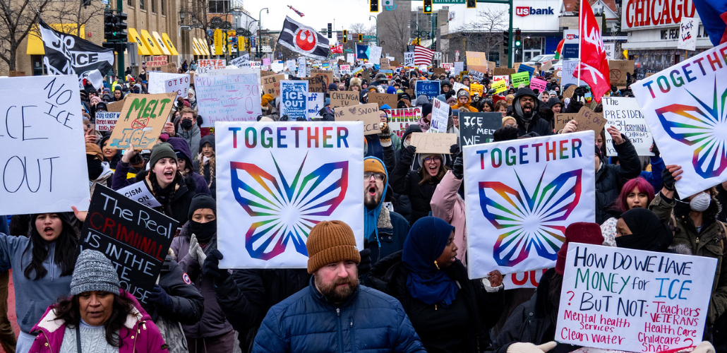 Mass march with many handmade anti-ICE signs. Most prominent are rainbow butterflies with the word "Together." Another in lower right says "How do have money for ICE but not teachers, veterans, childcare, health care, mental heath services, clean water, SNAP?" A flag farther back in crowd shows Star Wars rebellion logo.