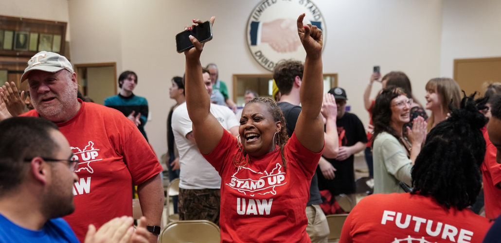 A Black woman in a red "Stand up UAW" T-shirt throws her hands in the air in celebration, phone in one hand, tears in her eyes. Other workers in matching shirts, and other people, stand nearby, all smiling, some clapping. In a room, looks like a union hall, with a logo on the wall that says "United we stand, divided we fall" around a handshake.