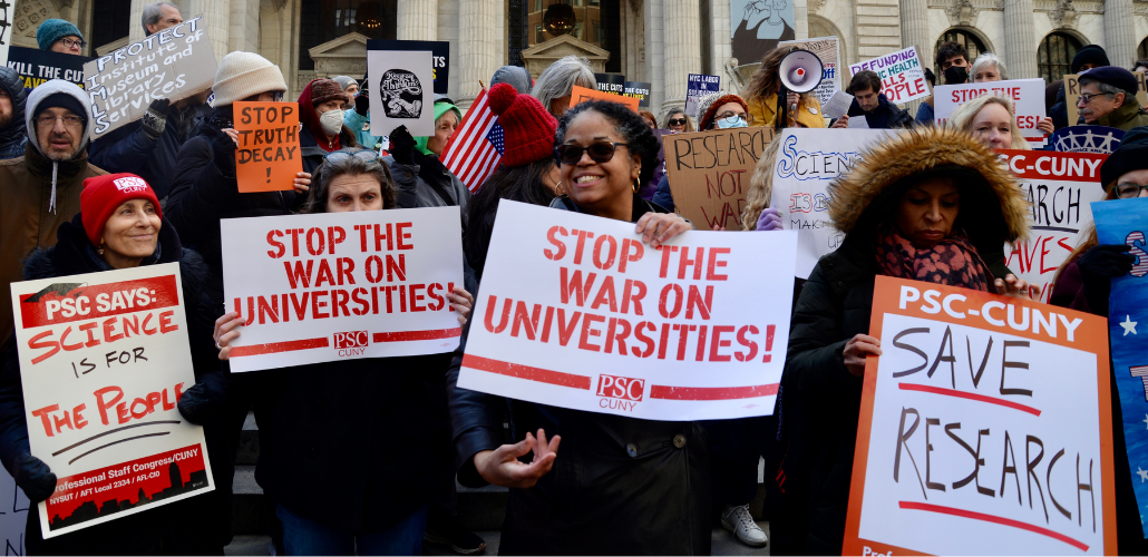 A big crowd, bundled against cold, stands in front of a building with columns. Foregrounded are four women, one with beaming smile, one looking up warmly at her. All hold PSC-CUNY signs that say variously, "Stop the War on Universities," "Save Research," and "Science is for the People." Other handmade signs, a UAW logo, and an upside-down American flag are visible in the crowd behind htem.