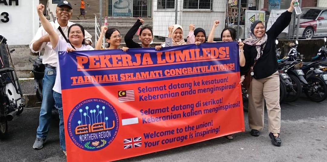A group of supporters stand behind a red and blue banner congratulating the Lumileds workers on voting for the union and saluting their courage.