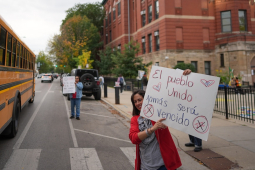 A teacher holds a sign between a school bus and the front of a school, with students visible in the background
