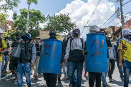 Row of protesters blockading the street in Colombia, two of whom are wielding blue shields of some kind.
