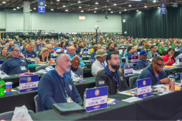 UAW delegates (men and women, Black and white) sit listening at the union's 2026 convention. Table signs identify those in the foreground as from Region 8, Locals 129 and 276.