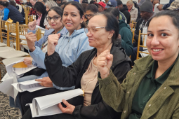 Four women of a range of ages, seated in a row, part of a bigger crowd in what looks like a hotel ballroom, smile and raise fists. Each holds in her lap a stapled packet of printed pages, perhaps a summary of a tentative agreement.