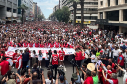 A huge crowd, as far as the eye can see, many wearking red, march down a street flanked by tall buildings.