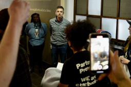 Two managers stand in a hallway as Starbucks workers gather around to confront them