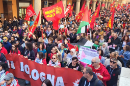 View of hundreds of people, a section of a giant march, filling an Italian street. Some marchers carry a huge red banner "FIOM-CGIL Bologna," some carry union flags or Palestinian flags, and one carries a handmade sign that says "Palestina Libera" with the flag and hearts. Mouths are open to sing or chant. Picturesque columns along the street. Marchers are diverse in race and gender.