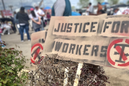 A hand-stenciled sign in the foreground says “Justice for Workers” while in the background striking workers stand outside a plant