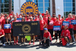 A group of 30 red-shirted autoworkers stand around a banner and hold contract campaign signs.