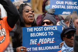 Two Black workers foregrounded in a dense crowd. Man on left wears orange New York Metro APWU T-shirt and lifts his hands to clap. Woman on right has thick eyelashes and holds a blue printed sign: "The Post Office belongs to the people, not the billionaires." Both smiling, energetic, probably chanting.