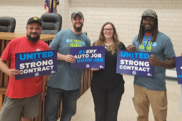 A group of five auto workers pose in their union hall with blue signs that say "End Tiers," "Every Auto Job a Good Job," and "United for a Strong Contract."