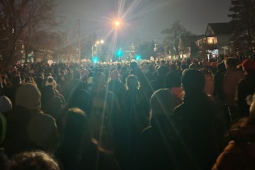 photo taken at night shows a giant crowd of people in a residential street, facing away from the viewer, silhouetted by a streetlight