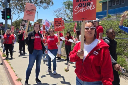 Teachers in red picket alongside a road with “Fully Staff Our Schools signs”