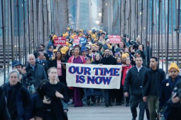 A dense and diverse crowd of people, some wearing yellow hats, marches across the Brooklyn Bridge in early-morning light. At the front are Zohran Mamdani and others carrying a banner that says in big purple letters "Our Time Is Now"