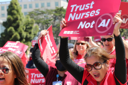 Women workers in red scrubs rally and chant, outdoors on a sunny day. They hold printed NNU signs. The woman most visible, in big sunglasses, holds a printed sign reading "Trust nurses, not A.I."