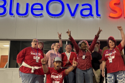 A group of 7 workers look excited and raise fists in front of a “BlueOval SK” sign