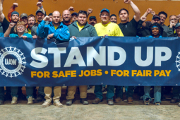 Thirty people, many with fists raised stand behind a banner that says “Stand Up, for safe jobs, for fair pay”
