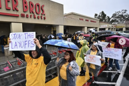 Workers in yellow shirts and rain parkas picket in front of a grocery store with a ‘stop the layoffs’ signs visible