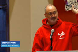 Screenshot from livestream. Shawn Fain in red "Liberators" sweatshirt stands at podium and holds up a copy of A Troublemaker's Handbook. Labels show his name, UAW President, and the livestream source: Act.tv.