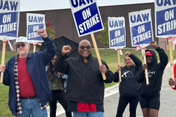 Six smiling workers carrying "UAW On Strike" picket signs raise their fists in the air outside a GM CCA facility. The workers are a mix of Black and white, men and women. Some of the picket signs are wrapped in plastic bags, and one worker carries an open umbrella.
