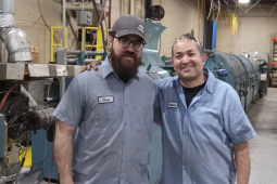 Two men in blue work shirts with nametags "Dan" and "Mohamed" sewn on stand side by side, arms around each other's shoulders, in a machine shop