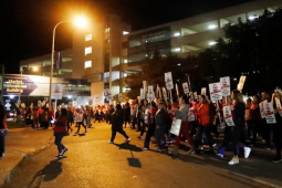Workers with "On Strike" picket signs march past a hospital in the near-dark, silhouetted by one bright light.