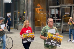Picketers march outside an office building, most in camouflage shirts. They carry signs: "UMWA ON STRIKE: Unfair Labor Practices"