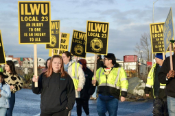 A group of union members and supporters march holding black and yellow ILWU Local 23 and Unfair Labor Practice signs.