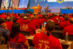 View from back of an auditorium at UAW convention. People in red shirts sit in rows; one red hoodie front and center has UAWD logo on the back. Onstage, Shawn Fain is at the podium, someone in a suit stands near him, and other people mostly in red shirts sit arrayed at long tables on the dais. A big blue screen behind them has UAW logo and intials repeated, and some other images not clearly visible.