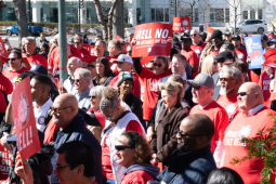A large crowd of people in red "Fight Like Hell" shirts stand outside in bright sunlight. Some hold printed NALC signs with the slogans "Hell No to Attacks on Us" and "Fight Like Hell!"
