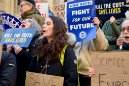 People, mostly white, rally outdoors on building steps. Printed signs say "Kill the cuts, save lives" and "UAW says: Fight for our future!" Handmade signs can't all be read but one says "Science belongs to everyone."