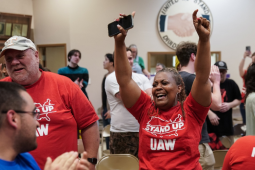A Black woman in a red "Stand up UAW" T-shirt throws her hands in the air in celebration, phone in one hand, tears in her eyes. Other workers in matching shirts, and other people, stand nearby, all smiling, some clapping. In a room, looks like a union hall, with a logo on the wall that says "United we stand, divided we fall" around a handshake. 