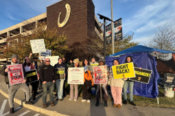 A dozen or so workers stand on a corner in from of a parking garage with a giant horseshoe logo on it. They look determined. They carry a mix of hand-lettered and printed signs saying things like "Dealers on strike," "Organize," "Fight back," "Horseshoe Teamsters on strike," and "On strike only against Horseshoe Casino Greed, Teamsters Local 135." Behind them is a canopy that has been insulated with tarps closing some of the sides. Camp chairs and a propane tank are visible. Some wear knitted hats.