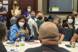 People are gathered around a table wearing masks, a woman with dark hair speaks and gestures in the middle.