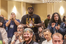 TDU convention attendees are shown sitting and standing, clapping for a speaker, in the hotel ballroom, under a chandelier.