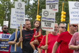 Auto workers march with signs that say “Stellantis Bargining in bad faith” One man is carrying a four-year old child.