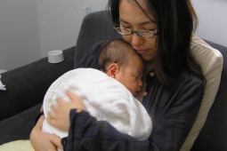 A seated woman with glasses in black holds a newborn baby wrapped in a white blanket to her chest.