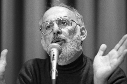 A bearded man speaks at a microphone with his hands wide open