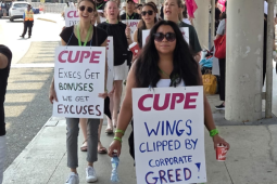 Flight attendants, mainly women, march on an airport sidewalk. Some wave pink CUPE flags and others wear sandwich-board signs with "CUPE" printed on and handwritten slogans. The two most visible say "Wings clipped by corporate greed!" and "Execs get bonuses, we get excuses." The woman in the foreground meets the camera's gaze with a serious expression; women behind her are chanting or singing.