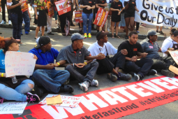 Fast food strikers, mostly young and Black, a mix of genders, sit in an intersection, linking arms. A long red banner laid out in front of them says "Whatever it takes." In the crowd behind them, some people hold a white handlettered banner: "Give us this day our daily bread."