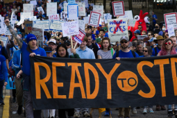 Big crowd marches behind a giant black banner hand-painted in orange "Ready to Strike." Many people hold handmade signs on sticks. A person near the front with long pink hair and reflecting sunglasses looks defiantly into the camera. A person in a blue beanie punches a tattooed arm into the air, raising a fist. Farther back, glimpse someone in a red inflatable lobster suit.