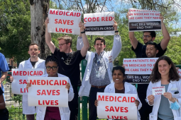 A diverse group of people, mostly in white lab coats, stand outside holding printed signs that say, in red on white, "Medicaid saves lives." Other signs say "Code red: patients in crisis" and "Our patients rely on Medicaid to get the care they need. This bill would end that."
