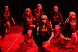 Seven women in long skirts and kerchiefs kneel, their arms in motion, apparently mid-dance. They are on a stage, bathed in red light.